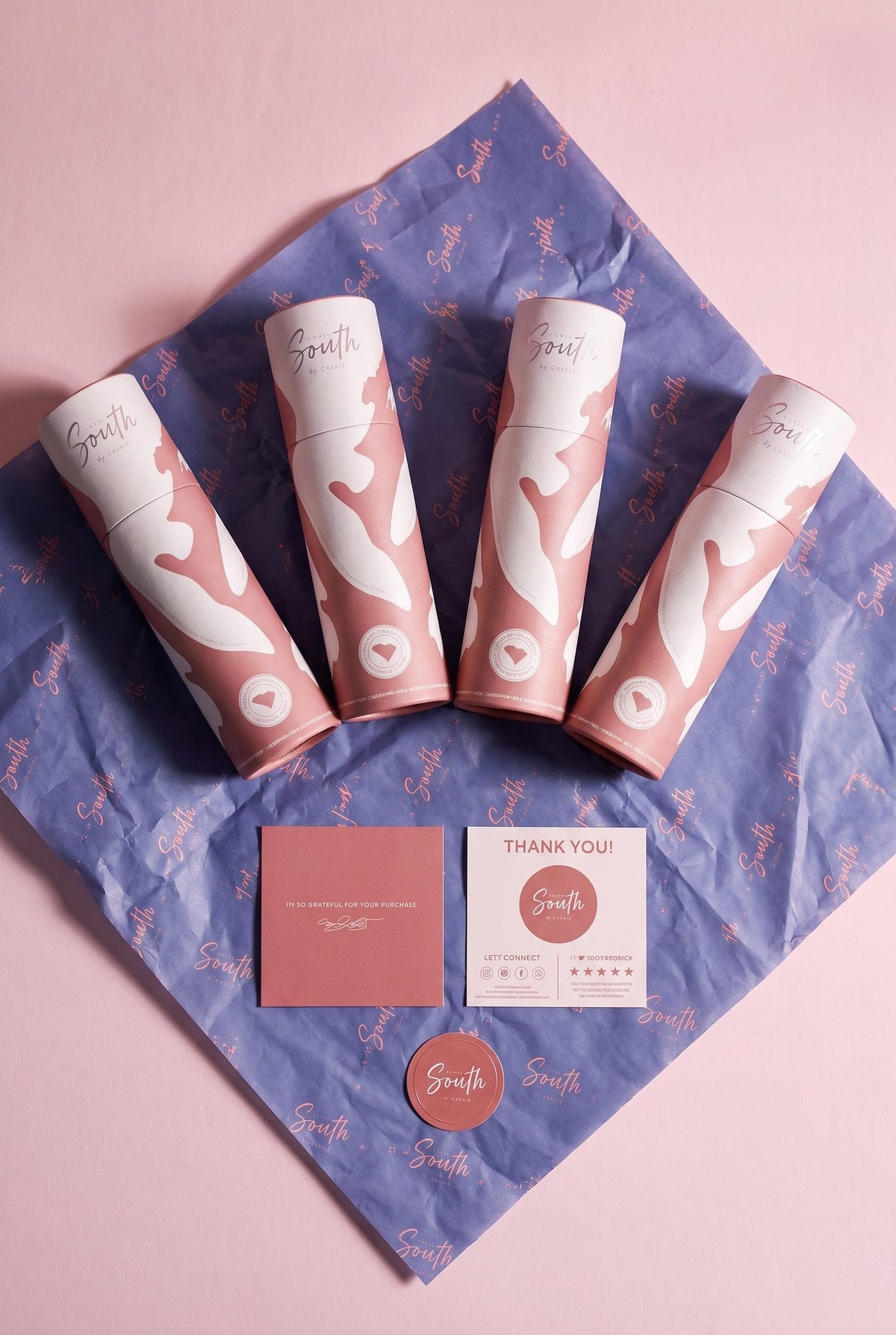 Design South art print and poster packaging with four coral branded shipping tubes arranged on purple tissue paper beside thank-you cards and a logo sticker.