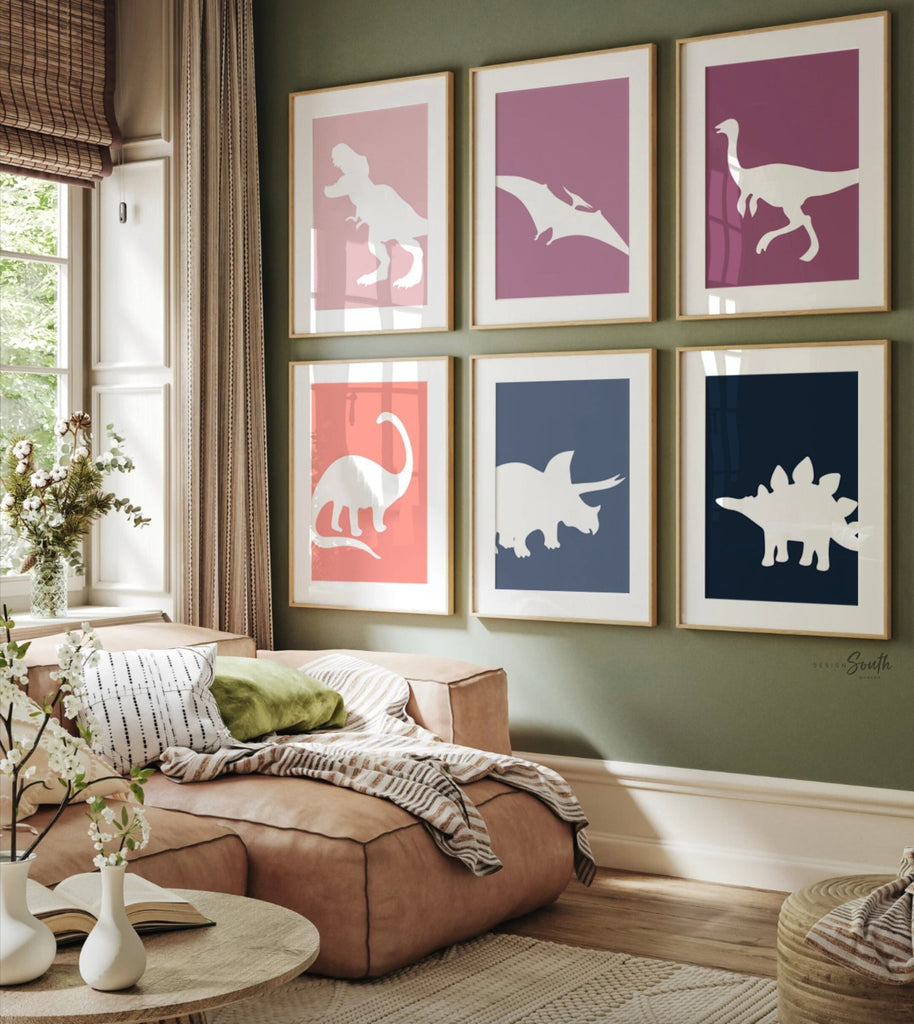 A set of six framed prints with white dinosaur silhouettes on colored backgrounds. The prints are arranged in two rows of three. The top row features a pink background with a T-Rex, a purple background with a Pterodactyl, and a pink background with a long-necked dinosaur. The bottom row features a pink background with a Brachiosaurus, a blue background with a Triceratops, and a blue background with a Stegosaurus. The prints are all framed in gold frames. The prints are hanging on a white wall.