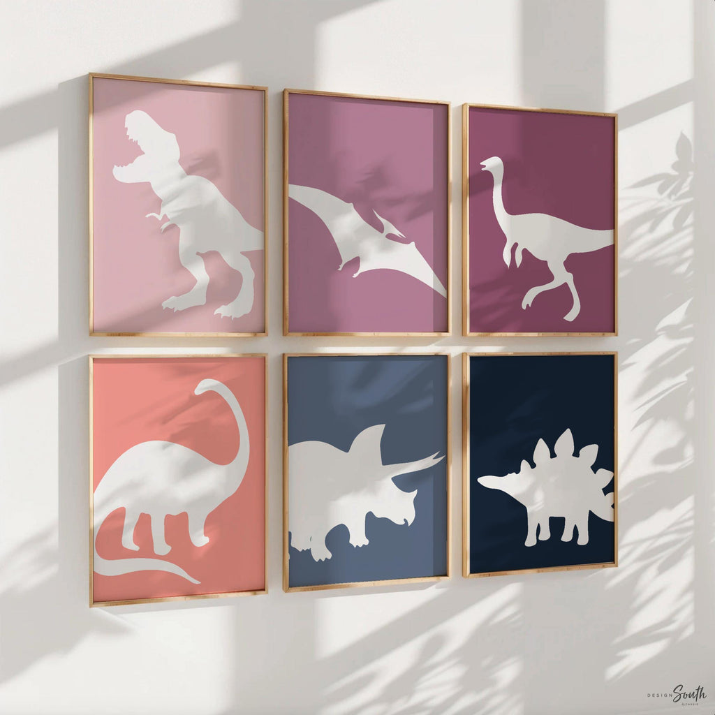 A set of six framed prints with white dinosaur silhouettes on colored backgrounds. The prints are arranged in two rows of three. The top row features a pink background with a T-Rex, a purple background with a Pterodactyl, and a pink background with a long-necked dinosaur. The bottom row features a pink background with a Brachiosaurus, a blue background with a Triceratops, and a blue background with a Stegosaurus. The prints are all framed in gold frames. The prints are hanging on a white wall.