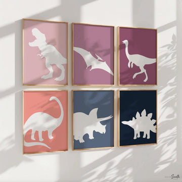 A set of six framed prints with white dinosaur silhouettes on colored backgrounds. The prints are arranged in two rows of three. The top row features a pink background with a T-Rex, a purple background with a Pterodactyl, and a pink background with a long-necked dinosaur. The bottom row features a pink background with a Brachiosaurus, a blue background with a Triceratops, and a blue background with a Stegosaurus. The prints are all framed in gold frames. The prints are hanging on a white wall.