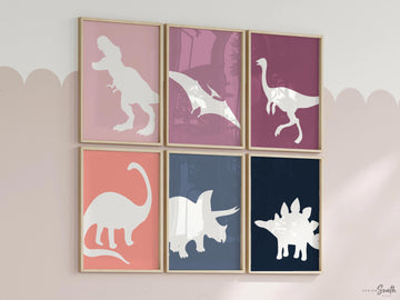 A set of six framed prints with white dinosaur silhouettes on colored backgrounds. The prints are arranged in two rows of three. The top row features a pink background with a T-Rex, a purple background with a Pterodactyl, and a pink background with a long-necked dinosaur. The bottom row features a pink background with a Brachiosaurus, a blue background with a Triceratops, and a blue background with a Stegosaurus. The prints are all framed in gold frames. The prints are hanging on a white wall.